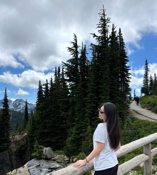 Photo of Flora standing in front of some trees, leaning on a fence and admiring the view. She is wearing a white shirt and sunglasses. 