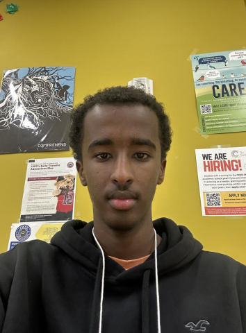 Photo of Yahya looking into the camera with a yellow wall and flyers behind him